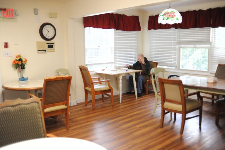 Senior resident sitting at a table in a well-lit dining area of an assisted living facility.