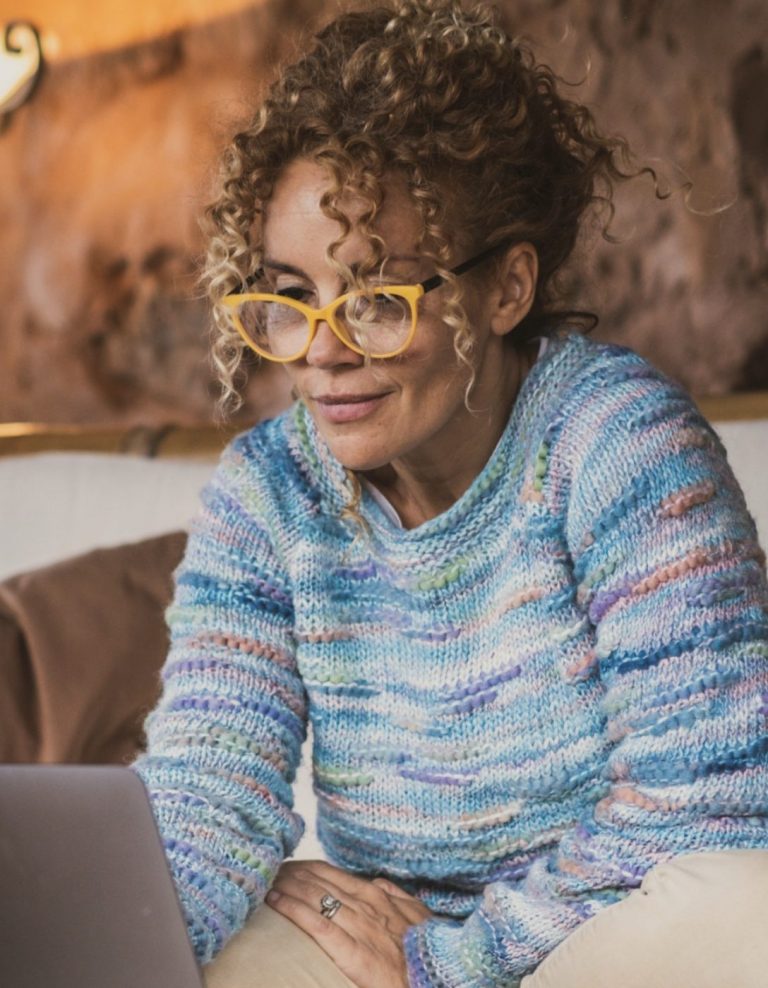 Woman with curly hair and yellow glasses using a laptop, wearing a cozy blue sweater, in a relaxed setting.